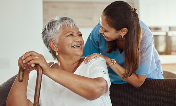 smiling elderly woman with gray hair and cane sitting on a couch while a younger woman stands behind her looking at her warmly indicating care and companionship in a comfortable setting with natural light suggesting harmony and joy in daily life with eight connections of love and support