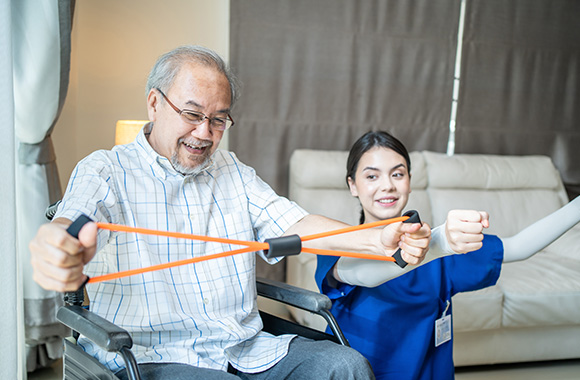 man in wheelchair using resistance band with female therapist in bright room focusing on rehabilitation and strength training techniques for seniors